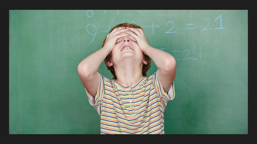 Ein Kind steht vor einer Schultafel und schaut nach oben. Das Kind schlägt die Hände über den Kopf zusammen, scheinbar verzweifelt von der Rechenaufgabe auf der Tafel. Was tun bei Dyskalkulie?
