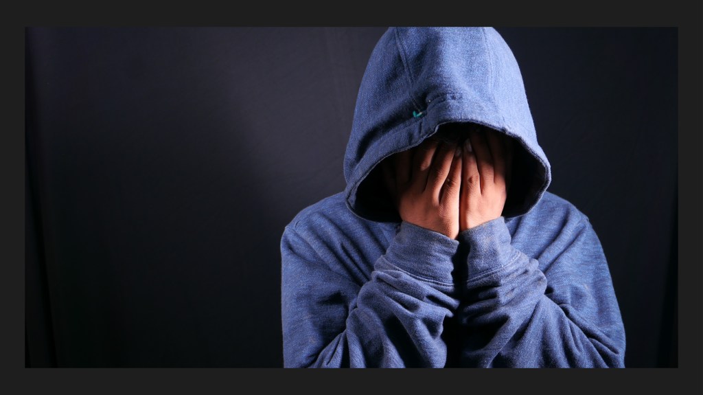 Ein Kind in einem dunklen Raum und allein hat sich eine Kapuze über den Kopf gezogen und die Hände vor das Gesicht geschlagen. Menschen mit Neurodivergenz fühlen sich oft wie Aliens in unserer Gesellschaft: allein und ausgeschlossen und ohne richtig kommunizieren zu können.