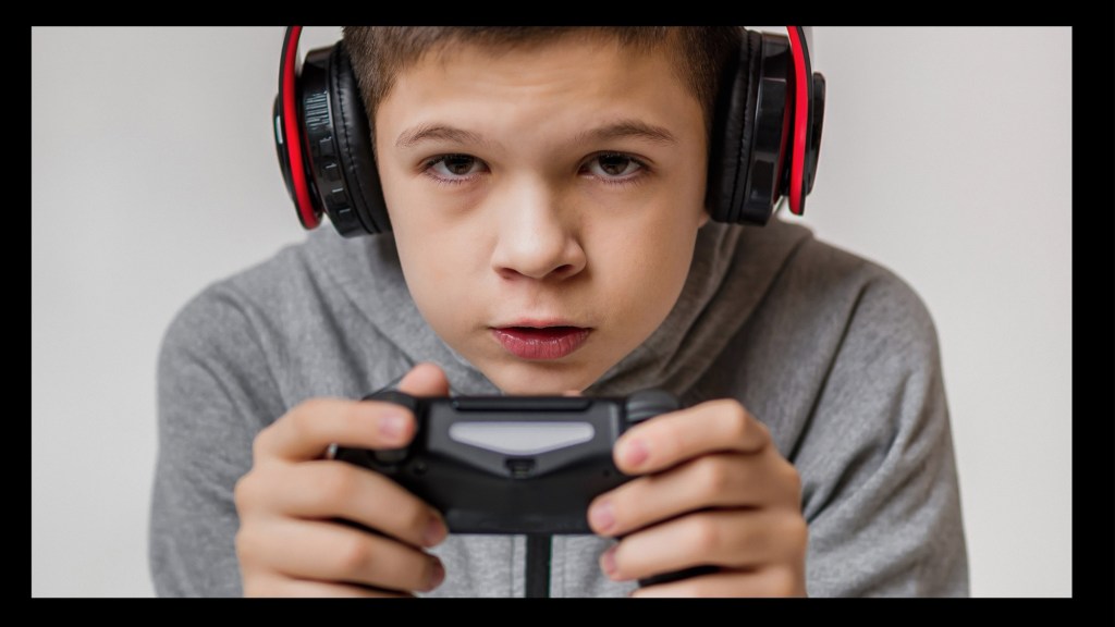 Ein Kind auf einem Bildschirm, den man nicht sehen kann, und hält eng umklammert einen Controller fest. Auf den Kopf hat es Kopfhörer: Warum zocken für Menschen mit Neurodiversität eine Gefahr ist.